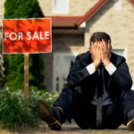 Man sitting near For Sale sign, holding his head.