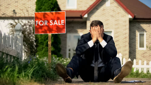 Man sitting near For Sale sign, holding his head.
