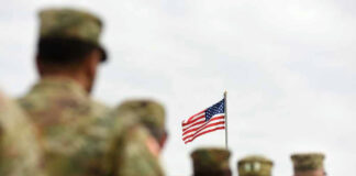 Soldiers in uniform standing, American flag in background.