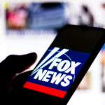WATCH: Fox News Guest’s Face Triggers WILD Speculation… Silhouette hand holding phone displaying Fox News logo.