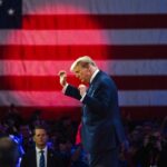 A speaker gesturing passionately in front of an American flag at a rally