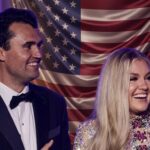 A smiling couple in formal attire standing in front of an American flag backdrop