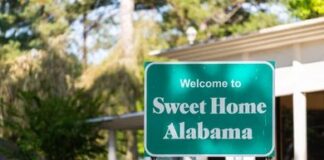 Green roadside sign reading Welcome to Sweet Home Alabama near a building and trees