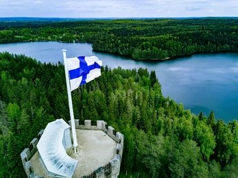 2140268127 Finnish flag flying on a stone tower above a forest and lake