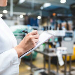 Person in lab coat writing on clipboard in factory.