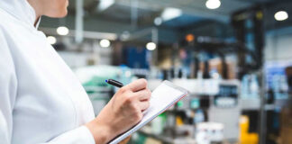 Person in lab coat writing on clipboard in factory.