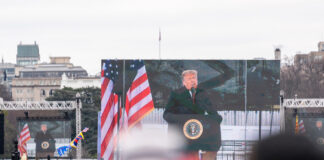 A large screen displays a speaker at a political rally with American flags in the foreground