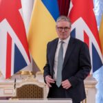 Man in suit stands before UK and Ukraine flags at a signing table