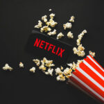 Spilled popcorn, phone displaying Netflix logo, striped popcorn container.