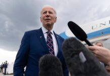 Man in blue suit speaking to microphones near airplane.