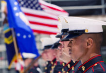 Marines in uniform stand at attention with American flag.