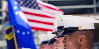 Marines in uniform stand at attention with American flag.