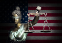 Blindfolded Lady Justice statue with scales, American flag backdrop.