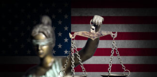 Blindfolded Lady Justice statue with scales, American flag backdrop.