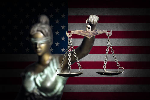 Blindfolded Lady Justice statue with scales, American flag backdrop.